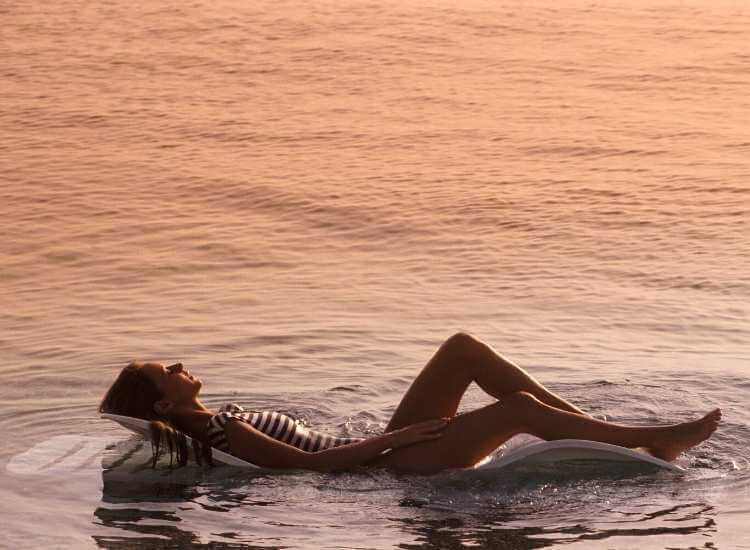 Mujer acostada en una silla de playa colocada dentro del mar, parcialmente sumergida, disfrutando del sol y la brisa marina.