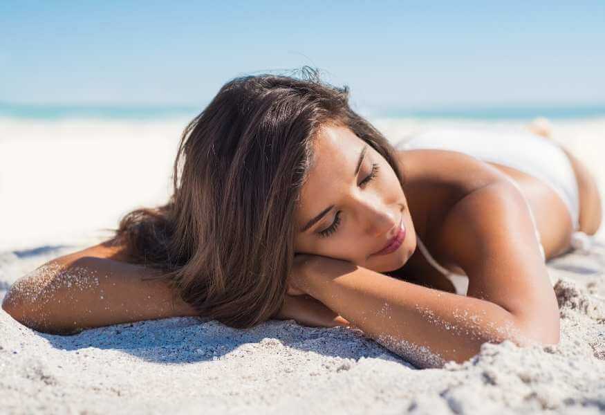 Mujer acostada boca abajo en la arena de una playa, exfoliando su piel y recibiendo brisa marina.